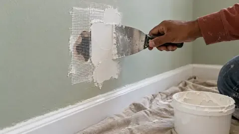 A putty knife spreading joint compound over a mesh patch covering a hole in a painted wall, with a tub of compound on a drop cloth